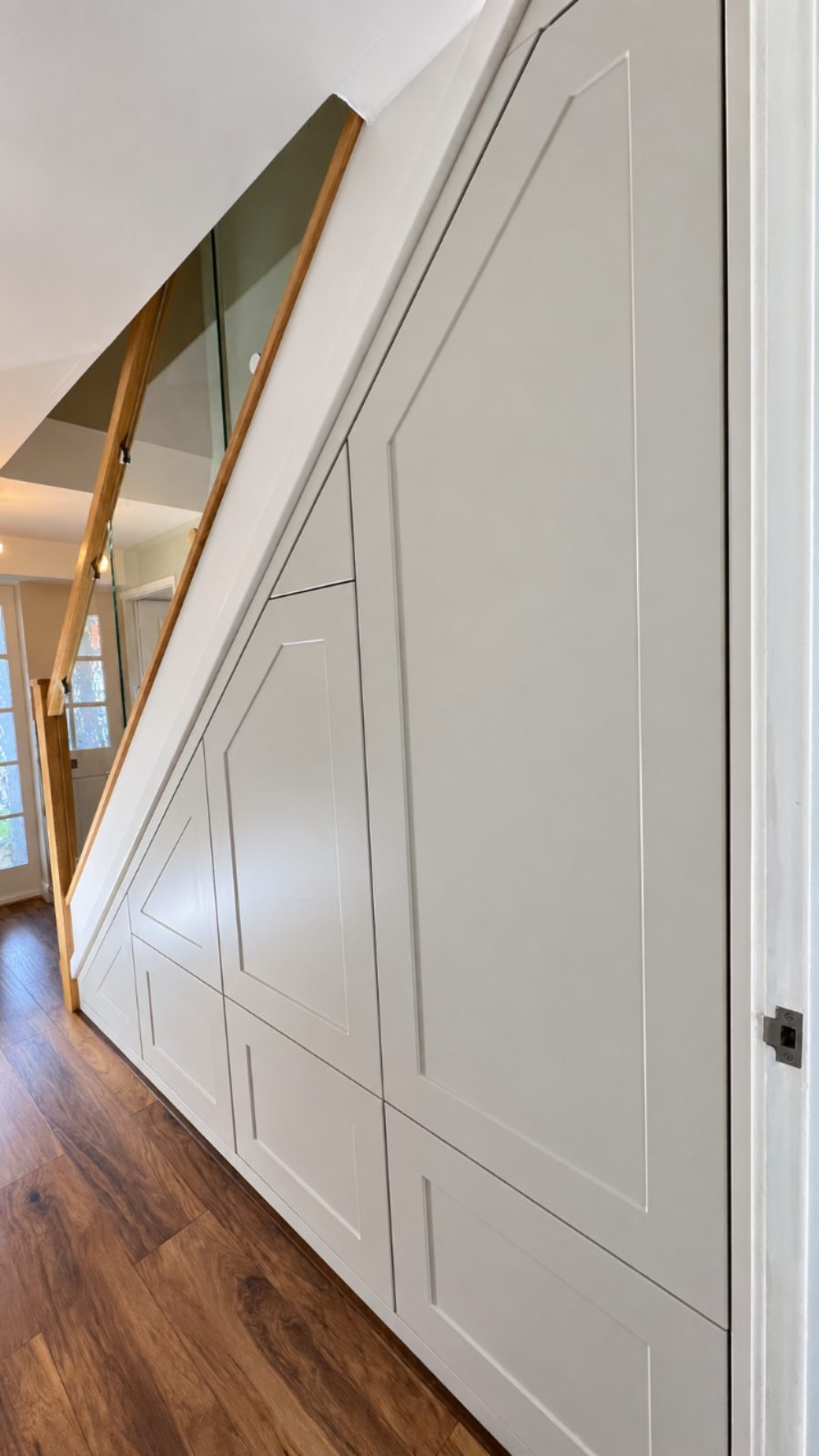 After: fitted understairs storage with five drawers and a larger cupboard in Porcelain Shaker fronts, following the line of the stairs, beneath a glass balustrade with an oak handrail